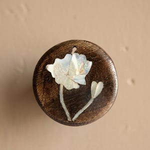 May include: A round, dark wooden pendant with a carved flower design. The flower is made of iridescent white shell pieces, with two stems and a bud. The pendant has a small hole at the top for hanging.