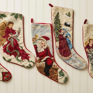 Könnte beinhalten: Fünf cremefarbene Weihnachtsstrümpfe aus Stoff mit detaillierten Stickereien von Weihnachtsmann, Engeln und Winterszenen. Jeder Strumpf hat einen roten Stoffbesatz an der Oberkante und eine kleine rote Schlaufe zum Aufhängen.