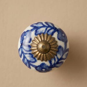 May include: Round ceramic cabinet knob with a white base and blue floral design. The knob features a brass-colored metal base and a central brass-colored screw. The knob is a decorative element for furniture.