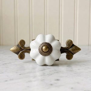 Könnte beinhalten: Ein Schubladengriff mit einem weißen, gewellten, blumenförmigen Keramikknopf, der auf einer dekorativen Rückplatte aus antikem Messing montiert ist. Die Rückplatte hat ein spitzes, von der Lilie inspiriertes Design und einen kleinen Messingniet.