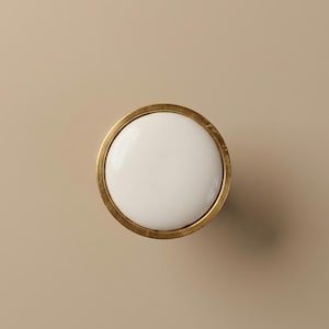 May include: A round cabinet knob with a white center and a gold-colored metal rim. The knob is centered against a neutral beige background, providing a clean and simple aesthetic. This hardware is suitable for various furniture styles.