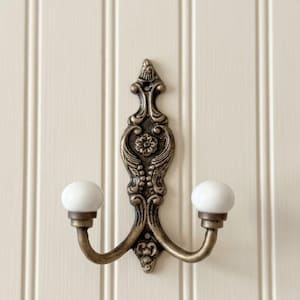 May include: A double wall hook featuring an antique bronze finish with ornate scrollwork and a central floral motif. The two curved arms extend outward, each ending in a smooth, round white ceramic knob for hanging items securely on the wall.