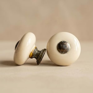 May include: Two off-white ceramic cabinet knobs with a dark metal base. One knob is upright, and the other is on its side, showing the metal base and the curvature of the knob. The knobs have a vintage or antique aesthetic.