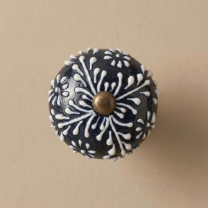 May include: A navy blue decorative cabinet knob with a white floral design. The knob has a brass-colored center and is round. The white floral pattern is raised, creating a textured surface. This decorative knob is suitable for cabinets and drawers.