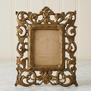May include: A rectangular picture frame with an ornate, metallic gold finish. The frame features intricate scrollwork and leaf-like patterns surrounding a central rectangular opening. The inner border has a textured, repeating geometric design.