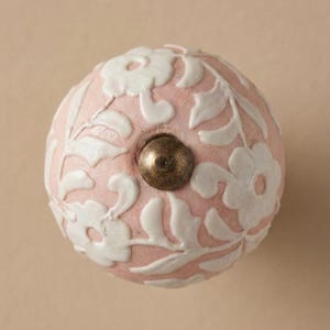 May include: A round, pink ceramic cabinet knob with a white floral design. The knob has a brass-coloured metal screw. The floral pattern is raised, creating a textured surface. This decorative knob is suitable for furniture.