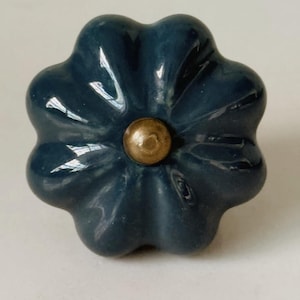 May include: A dark blue ceramic cabinet knob shaped like a six-petaled flower. The knob features a smooth, glossy finish and a centered, circular brass-toned metal accent piece that serves as the attachment point for the hardware.