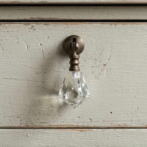 Op de afbeelding: Een hangende ladeknop met een heldere, gefacetteerde, diamantvormige glazen hanger bevestigd aan een ronde montageplaat van antiek bronskleurig metaal, gemonteerd op een lichtgrijze, verweerde houten ladefront.