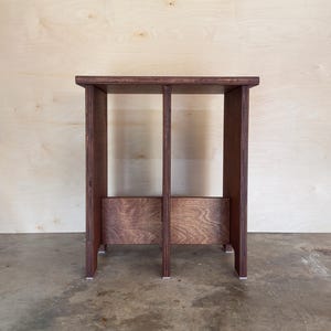 May include: A dark brown wooden stool with a rectangular top and two vertical supports. The stool has a simple, modern design and is set against a light wood background. The word "DROOPY" is printed in white at the bottom.