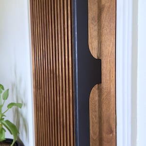 May include: A close-up of a wooden door with vertical slats and a black metal door handle. The handle has a unique design with a curved top and a straight bottom. The text "DROOPY" is visible at the bottom of the image.