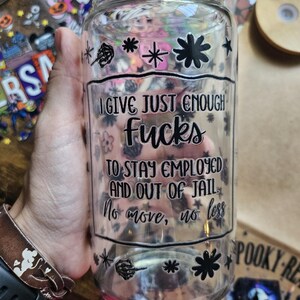 May include: A clear glass jar with a black graphic that reads "I GIVE JUST ENOUGH FUCKS TO STAY EMPLOYED AND OUT OF JAIL. No more, no less." The jar is decorated with black floral and hand-shaped designs.