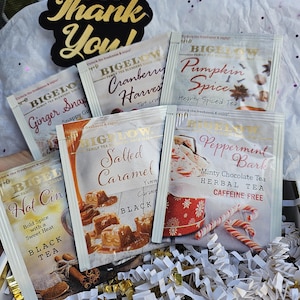May include: Assortment of Bigelow tea bags in various flavors, including Ginger Snap, Cranberry Harvest, Pumpkin Spice, Salted Caramel, and Peppermint Bark. The tea bags are arranged in a basket with shredded paper.