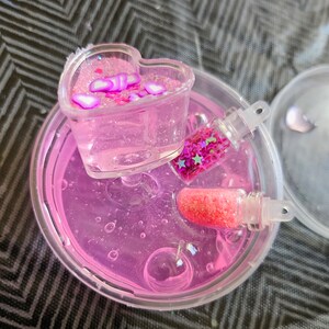 May include: A clear plastic container filled with pink slime, containing a heart-shaped container with pink and white glitter, and two small clear bottles with pink and white glitter.