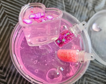 May include: A clear plastic container filled with pink slime, containing a heart-shaped container with pink and white glitter, and two small clear bottles with pink and white glitter.