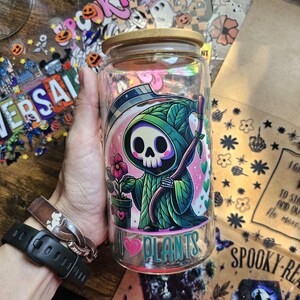 May include: A glass jar with a wooden lid features a cartoon image of the Grim Reaper holding a scythe and a potted plant. The jar has a pink heart with the text "I love plants" printed on it.