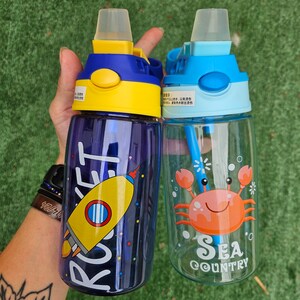 May include: Two colorful reusable water bottles with straw lids. The blue bottle has a cartoon crab with the text "Sea Country" and the purple bottle has a cartoon rocket with the text "Rocket".