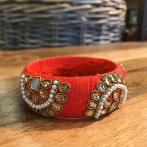 May include: An orange fabric bangle bracelet with gold and white embellishments. The bracelet is decorated with small, round, white pearls and gold-toned, faceted stones.