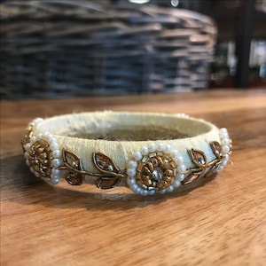 May include: A white fabric bangle bracelet with gold and pearl embellishments. The bracelet features a floral design with leaves and small flowers.