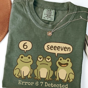 May include: Olive green t-shirt with a graphic of three cartoon frogs and the text "Error 6 7 Detected." The frogs are in speech bubbles that say "6" and "seeeven." A silver necklace with a small pendant is draped over the shirt.
