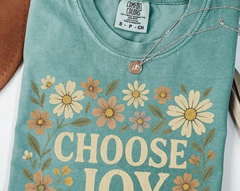 Op de afbeelding: Een turquoise Comfort Colors t-shirt met een bloemenontwerp en de woorden "CHOOSE JOY" in crème. Het shirt is versierd met een zilveren ketting. Het bloemenontwerp bevat madeliefjes en andere bloemen in crème, perzik en bruin.
