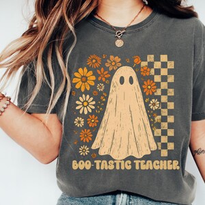 May include: Dark gray t-shirt featuring a ghost graphic with the text "Boo-tastic Teacher." The design includes orange and yellow flowers and a checkered pattern. The shirt is a comfortable, casual style.