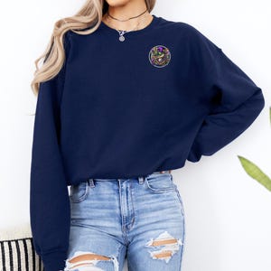 May include: A navy blue sweatshirt with a round, colorful graphic on the left chest. The graphic features a purple, green, and gold design.