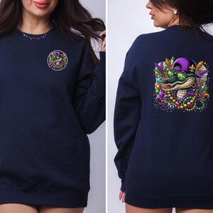 May include: A navy blue sweatshirt with a Mardi Gras alligator graphic on the back. The alligator is wearing sunglasses and a purple and gold hat. The front of the sweatshirt has a small, circular graphic of the alligator's head.