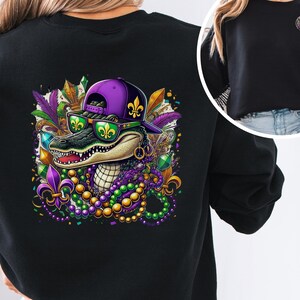 May include: Black sweatshirt with a colorful graphic of an alligator wearing a purple hat and sunglasses, surrounded by Mardi Gras beads and other decorations.