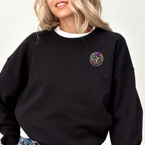 May include: Black sweatshirt with a round, colorful embroidered patch on the left chest. The patch features a jester's hat, beads, and other Mardi Gras-themed imagery.