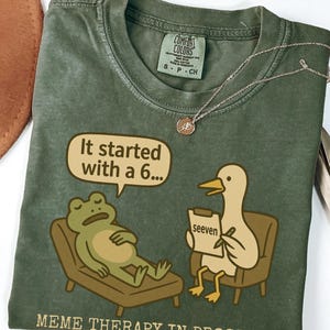 May include: Olive green t-shirt with a humorous meme design. The graphic features a frog on a therapist's couch and a duck with a notepad. Text includes "It started with a 6..." and "Meme Therapy in Progress."