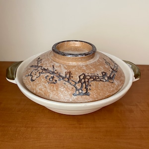 May include: A white ceramic pot with a brown lid and brown floral design. The pot has two green handles and is sitting on a wooden surface.