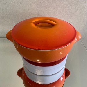 Könnte beinhalten: Ein orangefarbener und roter emaillierter Topf aus Gusseisen mit Deckel. Der Topf hat zwei Griffe an den Seiten und einen einzelnen Griff am Deckel.
