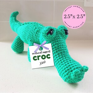 May include: Green crocheted crocodile stuffed animal with black eyes. The crocodile is holding a white tag that says "emotional support croc xoxo".