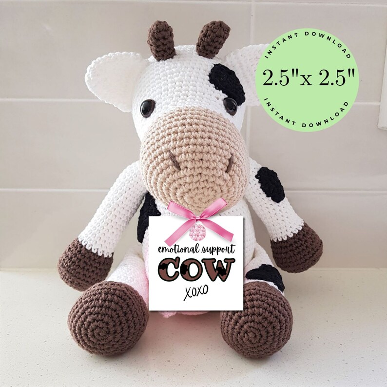 May include: A crocheted white cow plush toy with black spots, brown hooves, and a pink bow. The toy is holding a white tag that says "emotional support cow xoxo".
