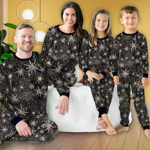 Könnte beinhalten: Passende Familien-Pyjama-Sets in Schwarz mit einem goldenen und weißen Feuerwerksmuster. Die langärmligen Oberteile und Hosen haben marineblaue Paspeln. Eine vierköpfige Familie trägt die Pyjamas und posiert auf einem weißen Kissen.