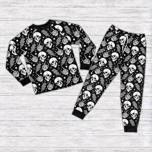 May include: Black pyjama set featuring a repeating pattern of white skulls, stars, and hand gestures on a black backdrop. The long-sleeved top and trousers have black cuffs and a black collar. The set is displayed on a white wood grain surface.