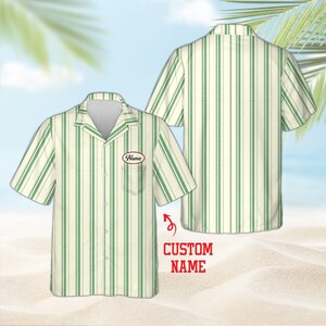 May include: A short-sleeved button-down shirt featuring a cream and green vertical striped pattern. The shirt includes a notched collar, a single chest pocket with an oval patch displaying the text Name, and a button-up front closure.