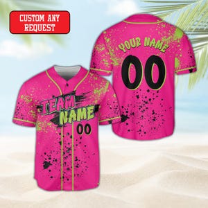 May include: A bright pink baseball jersey featuring a neon green and black paint splatter pattern. The front displays the text TEAM NAME and the number 00, while the back shows YOUR NAME and the number 00. A red box with CUSTOM ANY REQUEST is visible.