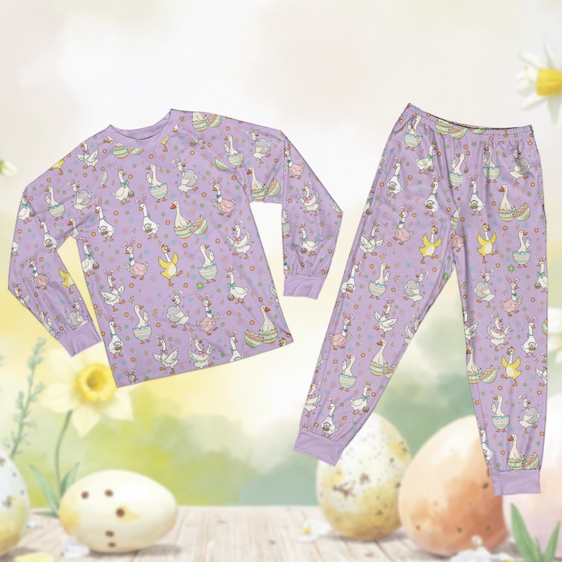 May include: A lavender pajama set featuring a repeating pattern of cartoon geese in Easter-themed outfits and eggs. The long-sleeved top and pants have lavender cuffs. The background includes daffodils and Easter eggs.