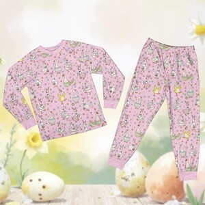 May include: Pink pajama set featuring a repeating pattern of geese, bunnies, and Easter eggs. The long-sleeved top and pants have pink cuffs. The whimsical design includes colorful accents and is perfect for Easter or spring.