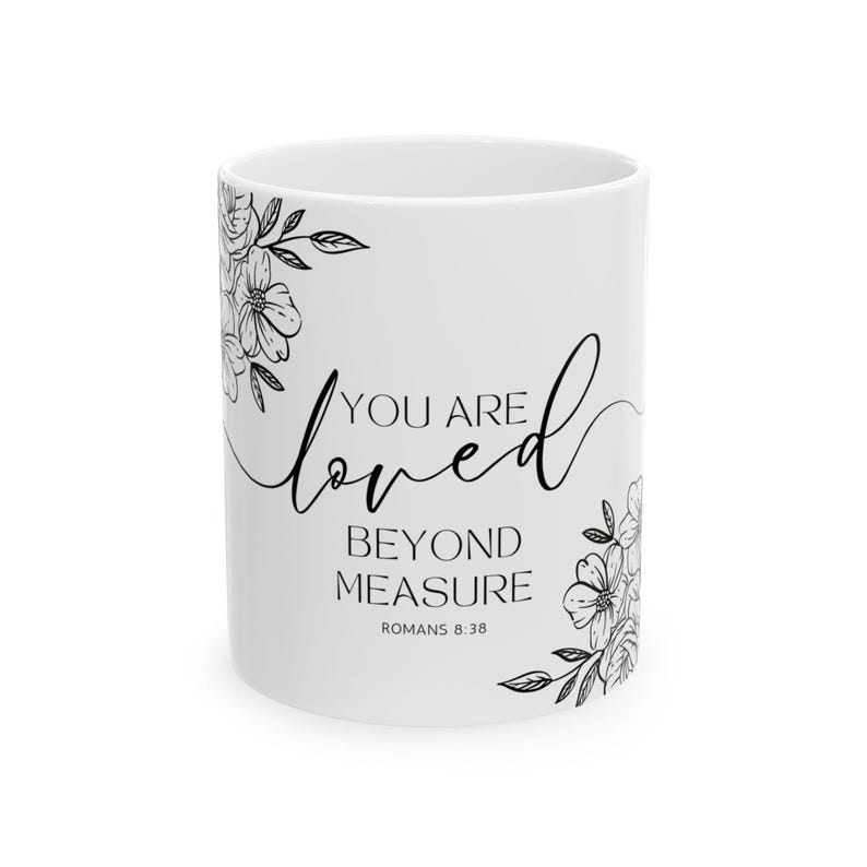 K&ouml;nnte beinhalten: Wei&szlig;e Keramiktasse mit schwarzem Blumendesign und dem Text "You are loved beyond measure Romans 8:38"