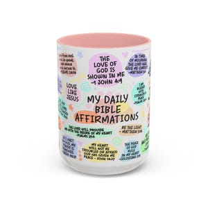 Op de afbeelding: Een witte keramische mok met een roze binnenkant, versierd met kleurrijke tekst en illustraties. De mok toont Bijbelse affirmaties, waaronder zinnen als "My Daily Bible Affirmations" en bijbelverwijzingen. Het ontwerp bevat pastelkleuren en een grillige stijl.