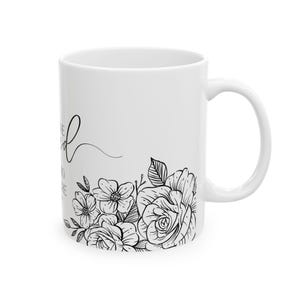 K&ouml;nnte beinhalten: Wei&szlig;e Keramiktasse mit schwarzem Blumendesign und dem Text "Sei nett zu dir selbst" in kursiv.