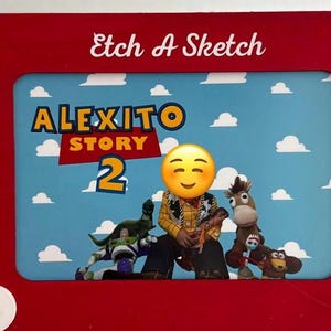 以下が含まれることがあります： 赤いエッチ・ア・スケッチのおもちゃ。背景は青色で、トイ・ストーリー2のキャラクターが描かれています。「Alexito Story 2」の文字が表示されています。おもちゃには白いノブと「Etch A Sketch」の文字が白で書かれています。