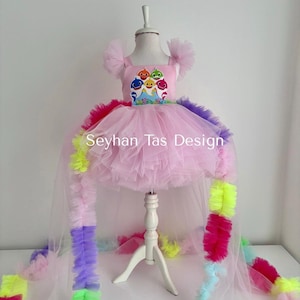 May include: Pink tulle dress with a Baby Shark graphic on the bodice. The dress features a high-low hemline and a long, rainbow-colored train. The text "Seyhan Tas Design" is visible on the front of the dress.