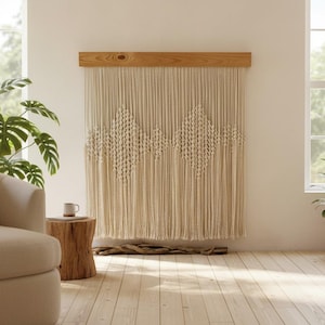 Può includere: Un grande arazzo in macramè color bianco sporco appeso a una trave di legno orizzontale. Il tessuto presenta complessi motivi a rombi annodati al centro di lunghi cordoni verticali che si estendono fino al bordo inferiore dell'oggetto.