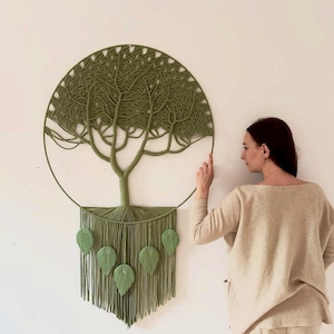 Puede incluir: Un gran tapiz circular de pared hecho de cuerda color verde salvia, con un intrincado diseño de árbol de macramé dentro de un aro. La sección inferior consta de flecos verticales adornados con cinco detalles de hojas de macramé.
