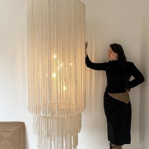 May include: A large, off-white macrame chandelier with a tiered design. The light fixture is cylindrical and hangs from the ceiling, with multiple light bulbs visible inside. The chandelier is made of woven, fringed material.