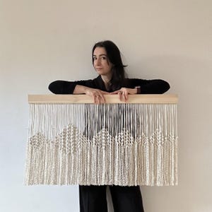 Puede incluir: Un tapiz de macramé blanco con un patrón geométrico y flecos. El tapiz está montado en un marco de madera.