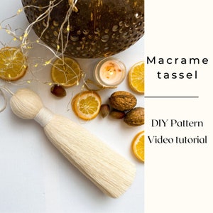 May include: A white macrame tassel with a DIY pattern and video tutorial. The tassel is hanging from a white surface with lemons, nuts, and a candle in the background.